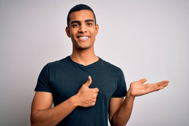 Junger gutaussehender afrikanisch-amerikanischer Mann in lässigem T-Shirt, das vor weißem Hintergrund steht Zeigt die Handfläche und macht eine ok Geste mit erhobenem Daumen, lächelt glücklich und fröhlich - Foto, Bild
