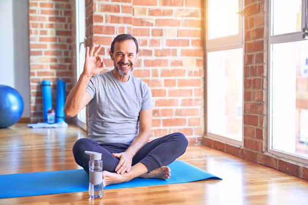Középkorú jóképű sportember ül szőnyegen csinál nyújtás jóga gyakorlat tornaterem mosoly pozitív csinál rendben jel kézzel és ujjakkal. Sikeres kifejezés. - Fotó, kép