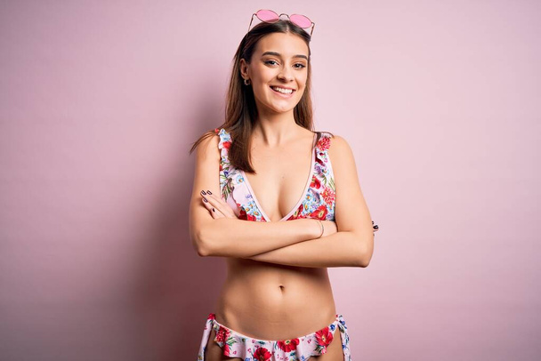 Jonge mooie vrouw op vakantie met bikini en zonnebril over roze achtergrond vrolijk gezicht glimlachend met gekruiste armen kijkend naar de camera. Positief persoon. - Foto, afbeelding