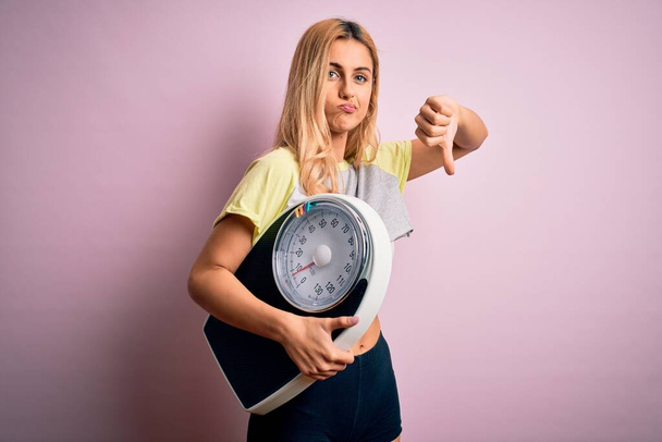 Молода красива блондинка спортивна жінка на дієті тримає вагу машини над рожевим тлом з розлюченим обличчям, негативний знак, що показує нелюбов з великими пальцями вниз, концепція відторгнення
 - Фото, зображення