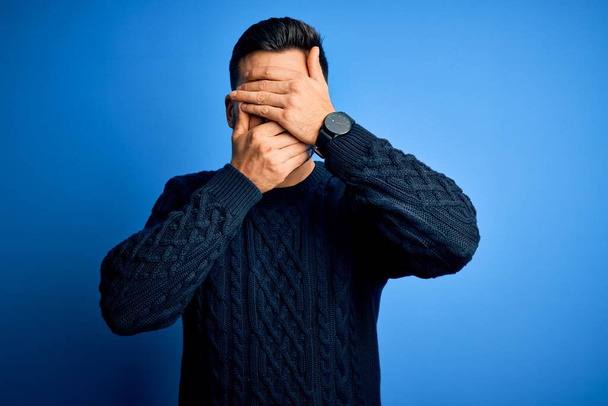 Young handsome man wearing casual sweater standing over isolated blue background Covering eyes and mouth with hands, surprised and shocked. Hiding emotion - Φωτογραφία, εικόνα