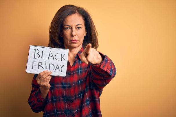 Femme brune du moyen âge tenant une publicité en papier noir vendredi sur fond isolé pointant du doigt la caméra et vers vous, signe de la main, geste positif et confiant de l'avant
 - Photo, image