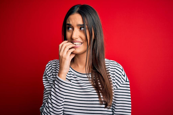 Young beautiful brunette woman wearing casual striped t-shirt over red background looking stressed and nervous with hands on mouth biting nails. Anxiety problem. - Foto, afbeelding
