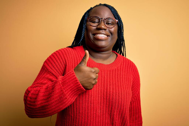 Mujer afroamericana de talla grande con trenzas que usan suéter casual sobre fondo amarillo haciendo un gesto feliz de pulgares hacia arriba con la mano. Aprobación de la expresión mirando a la cámara mostrando éxito
. - Foto, imagen