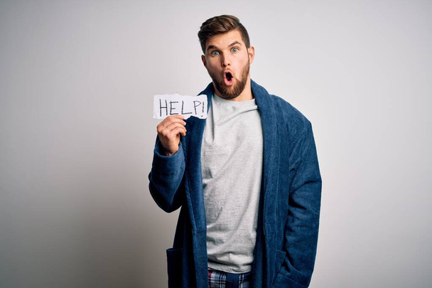 Young blond man with beard and blue eyes wearing pajama holding paper with help message scared in shock with a surprise face, afraid and excited with fear expression - Fotografie, Obrázek