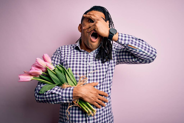 Giovane afroamericano afro romantico uomo con dreadlocks tenendo bouquet di tulipani rosa sbirciare in shock coprendo viso e occhi con la mano, guardando attraverso le dita con espressione imbarazzata
. - Foto, immagini