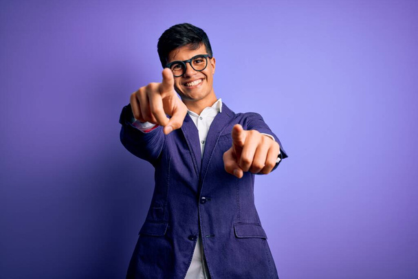 Молодой привлекательный бизнесмен в пиджаке и очках на изолированном фиолетовом фоне указывает пальцами на тебя и камеру, улыбаясь позитивно и весело
 - Фото, изображение