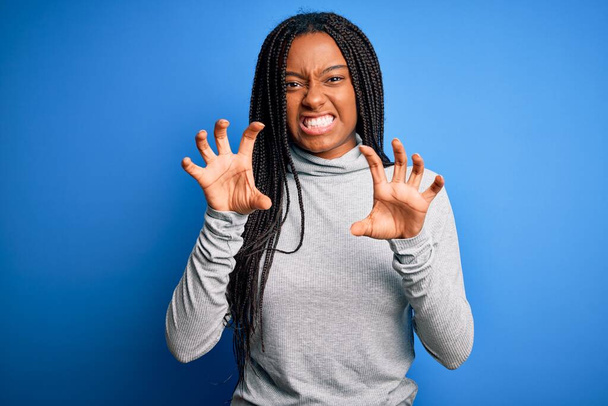 Junge afrikanisch-amerikanische Frau steht mit lässigem Rollkragen über blauem isoliertem Hintergrund und lächelt lustig und macht Krallen-Geste als Katze, aggressiver und sexy Ausdruck - Foto, Bild