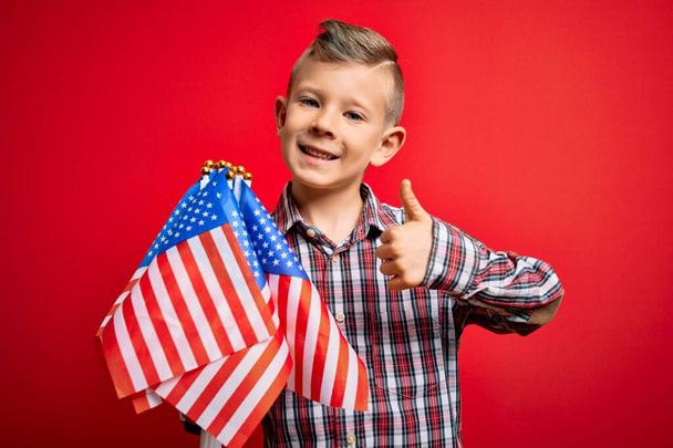 Junge kleine amerikanische patriotische kaukasische Kind hält Flagge der USA über rotem Hintergrund glücklich mit großem Lächeln tun ok Zeichen, Daumen nach oben mit den Fingern, hervorragendes Zeichen - Foto, Bild