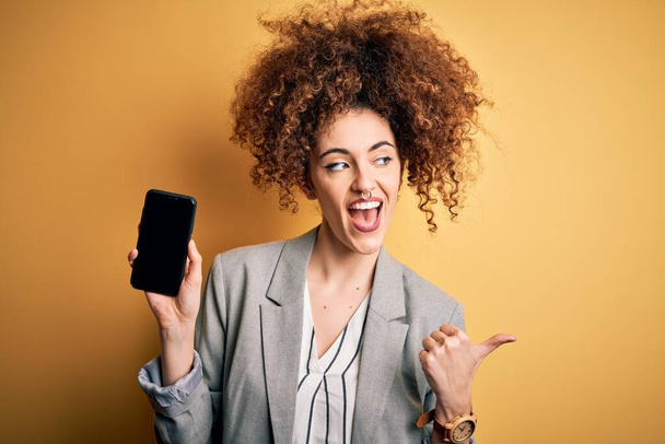 Молодая красивая женщина с вьющимися волосами и пирсинг держа смартфон показывая экран указывая и показывая с большим пальцем в сторону со счастливым лицом улыбаясь
 - Фото, изображение