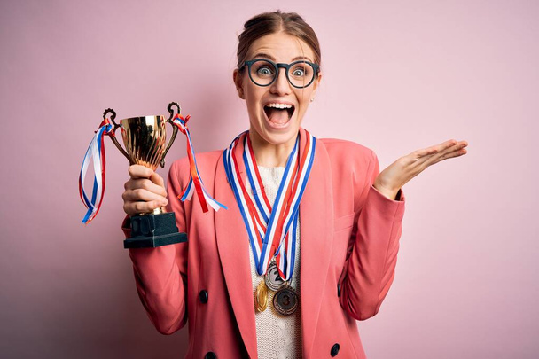 Young beautiful redhead woman holding trophy wearing medals over pink background very happy and excited, winner expression celebrating victory screaming with big smile and raised hands - Foto, Imagem