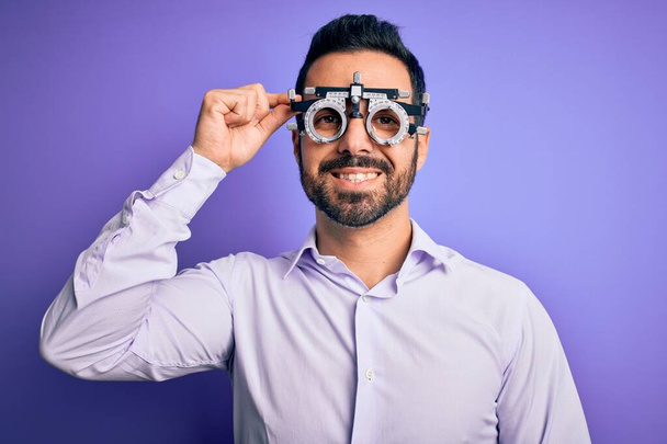 Young handsome optical man with beard wearing optometry glasses over purple background with a happy face standing and smiling with a confident smile showing teeth - Foto, Bild