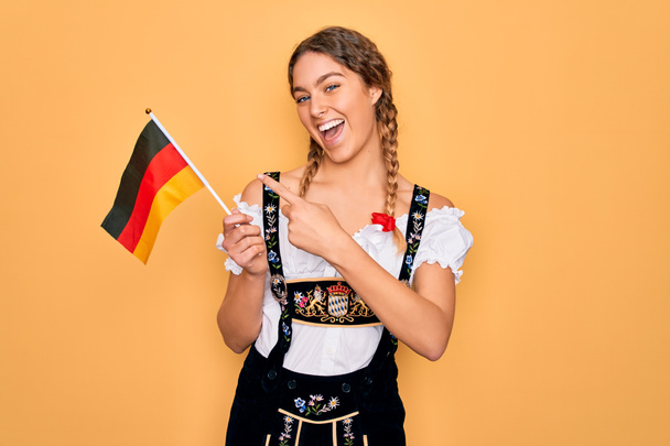 Linda mulher patriótica loira com olhos azuis vestindo vestido de octoberfest segurando bandeira alemã muito feliz apontando com mão e dedo
 - Foto, Imagem