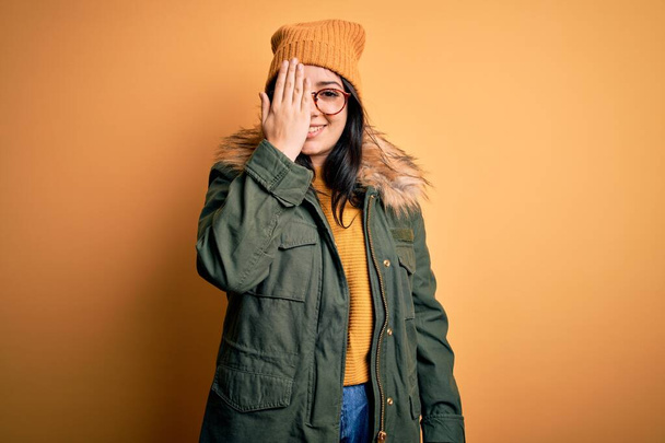 Młoda brunetka w okularach i zimowym płaszczu z kapeluszem na żółtym, izolowanym tle pokrywającym jedno oko ręką, pewny siebie uśmiech na twarzy i zaskakujące emocje. - Zdjęcie, obraz