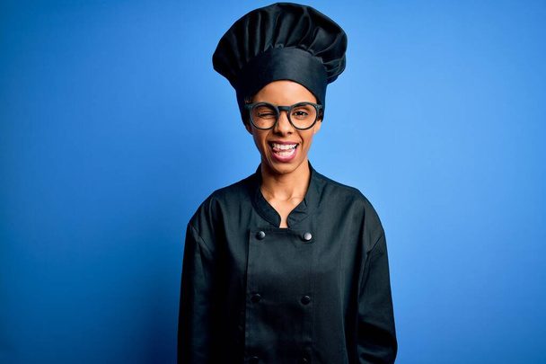 Young african american chef woman wearing cooker uniform and hat over blue background winking looking at the camera with sexy expression, cheerful and happy face. - Φωτογραφία, εικόνα
