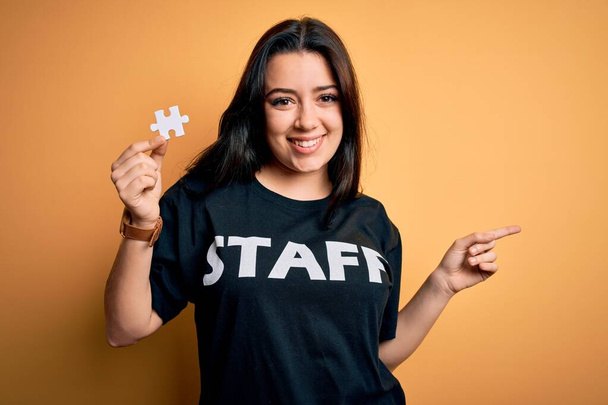 Young brunette worker woman wearing staff t-shirt as uniform showing puzzle piece as teamwork very happy pointing with hand and finger to the side - Fotó, kép