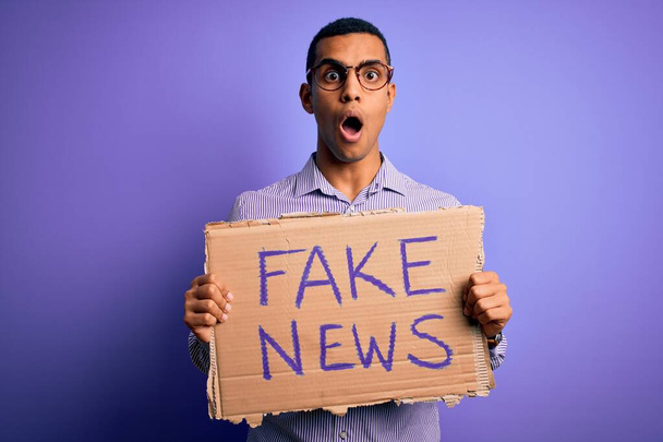 Junger gut aussehender afrikanisch-amerikanischer Mann protestiert mit einem Transparent mit Fake News Botschaft, erschrocken mit einem überraschten Gesicht, ängstlich und aufgeregt mit Angstausdruck - Foto, Bild