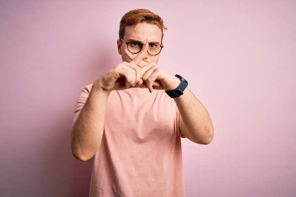 Young handsome redhead man wearing casual t-shirt standing over isolated pink background Rejection expression crossing fingers doing negative sign - Fotografie, Obrázek