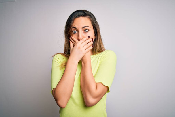 Beautiful blonde woman with blue eyes wearing green casual t-shirt over white background shocked covering mouth with hands for mistake. Secret concept. - Foto, afbeelding