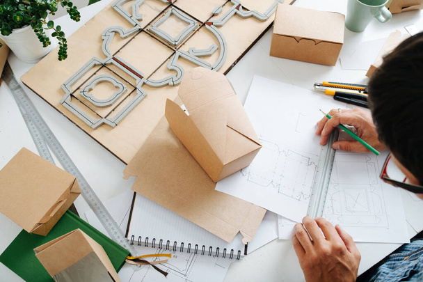 Top view of workplace packaging designer. A man work on the table with drawings, rulers and pencils, ready-made boxes and punching platen - Photo, image