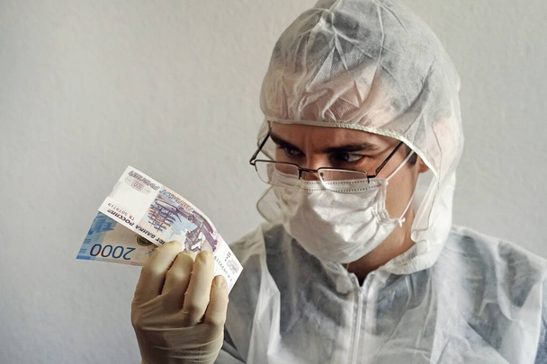 Koruyucu kıyafetli, tıbbi eldivenli ve maskeli bir bilim adamı elinde nakit para tutuyor. Kirli mikroplar ve para üzerindeki virüs bakterileri. - Fotoğraf, Görsel