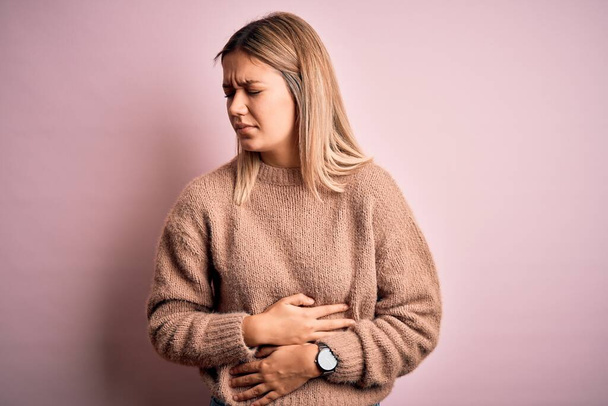 Jonge mooie blonde vrouw dragen winterwol trui over roze geïsoleerde achtergrond met de hand op de maag omdat indigestie, pijnlijke ziekte gevoel onwel. Ache-concept. - Foto, afbeelding