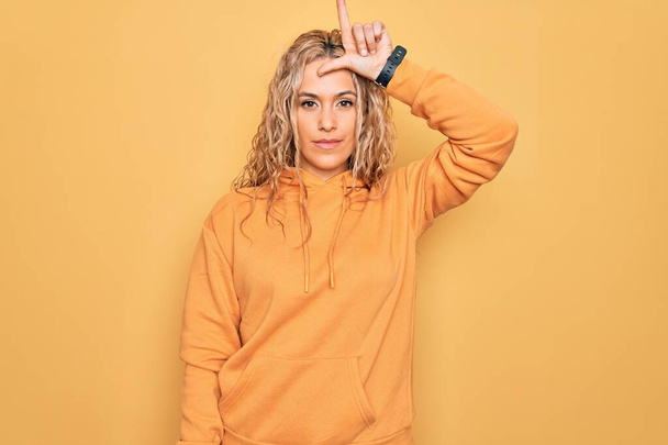 Jeune belle femme sportive blonde portant un sweat-shirt décontracté sur fond jaune se moquant des gens avec les doigts sur le front faisant un geste de perdant moqueur et insultant
. - Photo, image