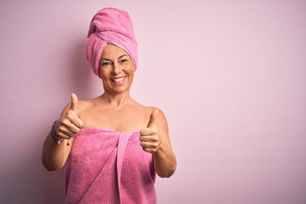 Жінка середнього віку носить рушник для ванни від догляду за тілом краси на рожевому тлі, схвалюючи позитивний жест рукою, великі пальці посміхаються і щасливі за успіх. жест переможця
. - Фото, зображення