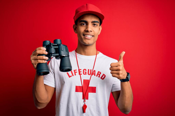 Young handsome african american lifeguard man wearing whistle using binoculars happy with big smile doing ok sign, thumb up with fingers, excellent sign - Photo, image