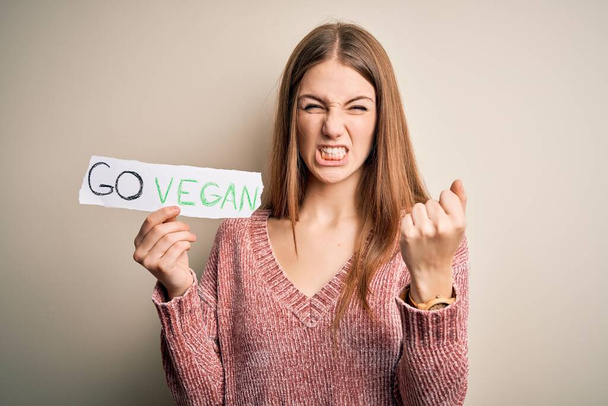 Νεαρή όμορφη κοκκινομάλλα γυναίκα ζητά veggie τροφίμων κρατώντας χαρτί με vegan μήνυμα ενοχλημένος και απογοητευμένος φωνάζοντας με θυμό, τρελός και φωνάζοντας με σηκωμένο χέρι, έννοια του θυμού - Φωτογραφία, εικόνα