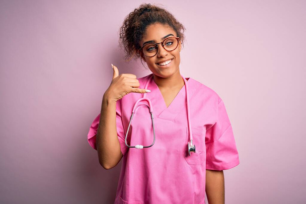 ピンクの背景に医療用の制服を着たアフリカ系アメリカ人看護婦の少女が、電話で話すような手や指で携帯電話のジェスチャーをして笑っている。概念の伝達. - 写真・画像