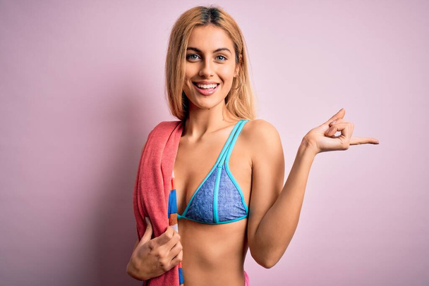 Hermosa mujer rubia de vacaciones con bikini sosteniendo toalla de playa sobre fondo rosa muy feliz señalando con la mano y el dedo a un lado
 - Foto, imagen