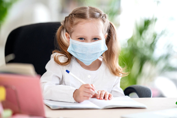 Cute little girl, doing homework, writing in a notebook , using a laptop, digital e -learning concepts, during quarantine and self- isolation. Remote e-learning due to Covid-19 coronavirus pandemic - Fotografie, Obrázek