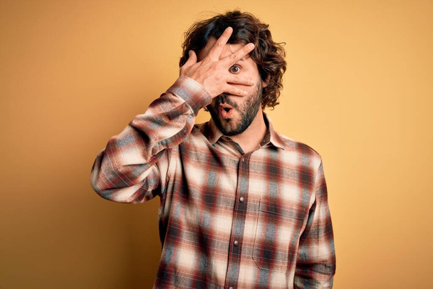 Mladý pohledný muž s vousy v neformální košili stojící nad žlutým pozadím vykukující v šoku a zakrývající obličej a oči rukou, dívající se skrz prsty s rozpačitým výrazem. - Fotografie, Obrázek