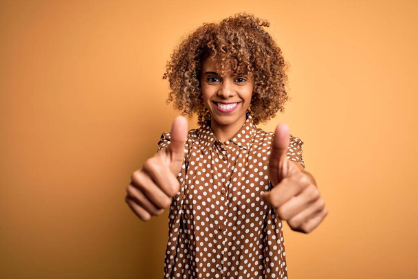 A fiatal, gyönyörű afro-amerikai nő alkalmi inget visel, sárga háttér felett áll, és pozitív gesztust tesz kézzel, hüvelykujjával mosolyogva örül a sikernek. Győztes gesztus. - Fotó, kép