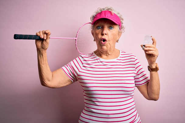 Senior schöne Sportlerin hält Badmintonschläger über isolierten rosa Hintergrund erschrocken mit einem Überraschungsgesicht, Angst und aufgeregt mit Angst Ausdruck - Foto, Bild