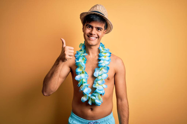 Jeune homme touriste beau en vacances portant des maillots de bain et des fleurs hawaïennes de lei faisant geste heureux pouces vers le haut avec la main. Approuver l'expression en regardant la caméra montrant le succès. - Photo, image