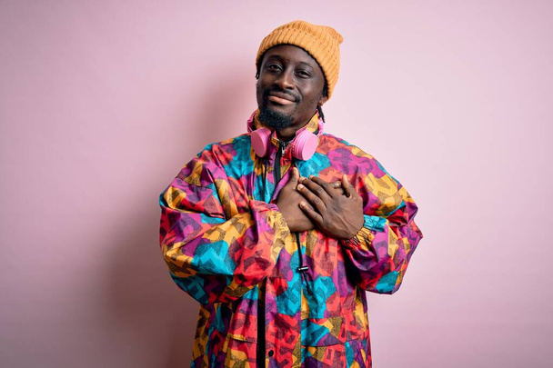 Νεαρός όμορφος Αφροαμερικάνος που φοράει πολύχρωμο παλτό και καπέλο πάνω από ροζ φόντο χαμογελώντας με τα χέρια στο στήθος με κλειστά μάτια και ευγνώμων χειρονομία στο πρόσωπο. Έννοια υγείας. - Φωτογραφία, εικόνα