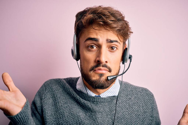 Młody agent call center z brodą, noszący słuchawki na odizolowanym różowym tle, nieświadomy i zdezorientowany wyraz twarzy z podniesionymi rękami. Koncepcja wątpliwości. - Zdjęcie, obraz