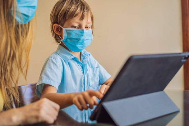 Boy studying online at home using a tablet. Mom helps him learn. Mom and son in medical masks to protect against coronovirus. Studying during quarantine. Global pandemic covid19 virus - 写真・画像