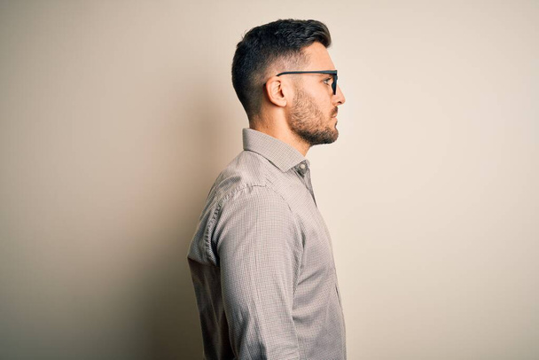 Junger gutaussehender Mann mit elegantem Hemd und Brille über isoliertem weißem Hintergrund, der zur Seite schaut, entspannte Profil-Pose mit natürlichem Gesicht mit selbstbewusstem Lächeln. - Foto, Bild
