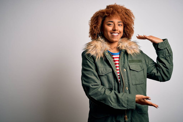 Young beautiful African American woman with curly hair wearing green casual winter coat gesturing with hands showing big and large size sign, measure symbol. Smiling looking at the camera. Measuring concept. - Photo, Image