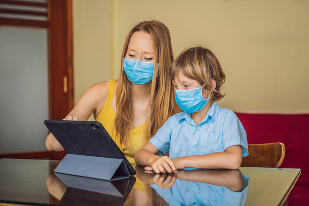 Boy studying online at home using a tablet. Mom helps him learn. Mom and son in medical masks to protect against coronovirus. Studying during quarantine. Global pandemic covid19 virus - Photo, image