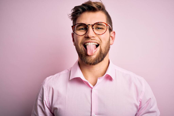 Joven hombre rubio guapo con barba y ojos azules con camisa rosa y gafas que salen con la lengua feliz con expresión divertida. Concepto de emoción
. - Foto, imagen