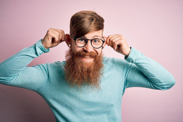 Przystojny irlandzki rudzielec z brodą w okularach na różowym, odizolowanym tle Uśmiechnięty, ciągnący uszy palcami, zabawny gest. Problem przesłuchań - Zdjęcie, obraz