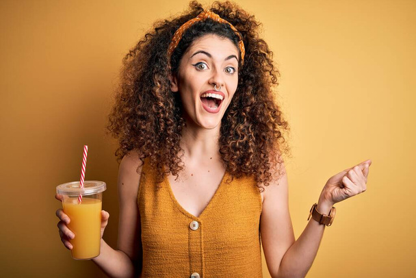Jeune belle femme aux cheveux bouclés et perçant boire du jus d'orange sain criant fière et célébrant la victoire et le succès très excité, acclamant l'émotion
 - Photo, image