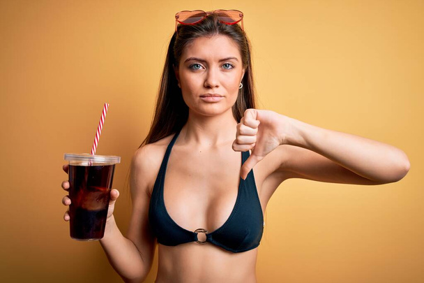 Young beautiful woman with blue eyes on vacation wearing bikini drinking cola beverage with angry face, negative sign showing dislike with thumbs down, rejection concept - Photo, Image