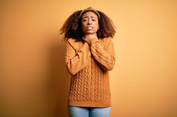 Mujer afroamericana joven con el pelo afro usando suéter casual sobre fondo amarillo gritando y sofocándose porque estrangulamiento doloroso. Problema de salud. Concepto de asfixia y suicidio
. - Foto, imagen