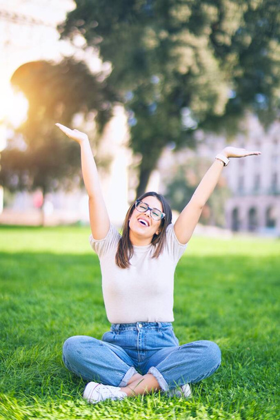 Młoda piękna kobieta uśmiechnięta szczęśliwa i pewna siebie. Siedząc na trawie z uśmiechem na twarzy w parku - Zdjęcie, obraz