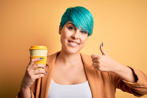 Nuori nainen sininen muoti hiukset juominen kuppi kahvia keltainen eristetty tausta onnellinen iso hymy tekee ok merkki, peukalo ylös sormilla, erinomainen merkki
 - Valokuva, kuva